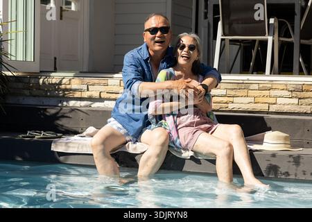 Senior couple sitting on cushioned pool ledge laughing, wearing sunglasses near straw hat Stock Photo