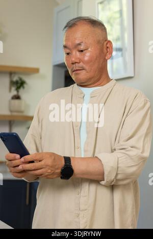 Senior Asian man standing in kitchen, checking smartphone, wearing beige shirt and black wristwatch Stock Photo