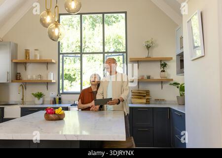 Senior Asian couple standing in kitchen behind marble island holding tablet and looking upward Stock Photo