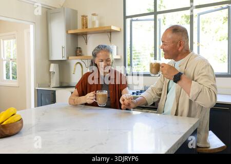 Senior Asian couple smiling and holding hands standing at kitchen island with glass coffee mugs Stock Photo