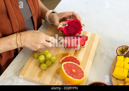 Paring knife slicing magenta dragon fruit on wood board on marble counter, mango, passionfruit Stock Photo