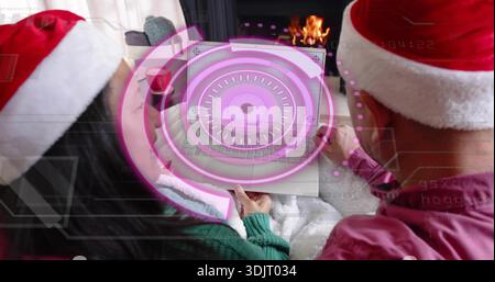 Sharing couple wearing Santa hats, viewing laptop pink hologram at home, white blanket, fireplace Stock Photo