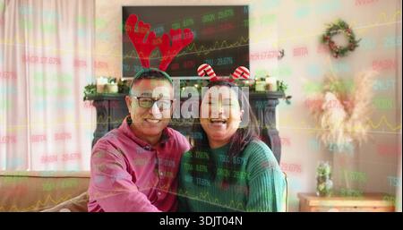 Laughing couple wearing novelty headbands and sweaters on sofa at home with stock market overlay Stock Photo