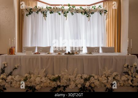 A beautifully arranged wedding head table with white floral garlands, candlesticks, and fine dinnerware set against sheer curtains and golden drapery Stock Photo