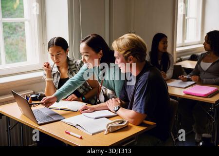 Female professor using laptop while assisting students sitting at desk in classroom at college Stock Photo