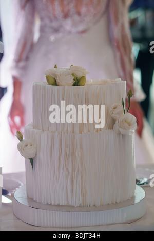 A beautifully crafted white ruffled wedding cake adorned with delicate white roses and green buds, displayed in front of a bride in a lace gown, captu Stock Photo