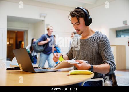 Young man wearing wireless headphones studying while sitting at table in college campus Stock Photo