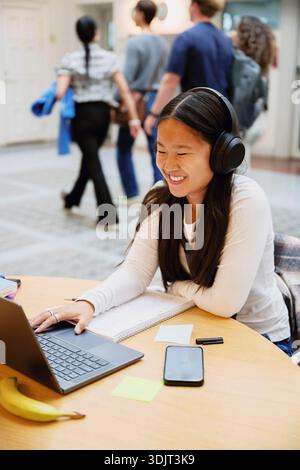 Smiling young woman wearing wireless headphones using laptop while sitting at table in university campus Stock Photo