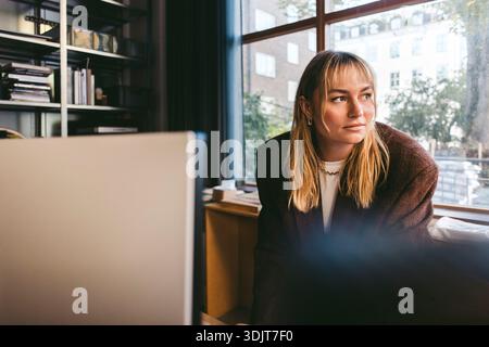 Young female creative professional looking away while leaning on desk in office Stock Photo