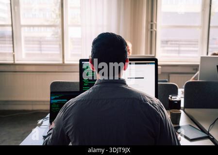 Rear view of young male programmer working on computers while sitting at desk in tech office Stock Photo