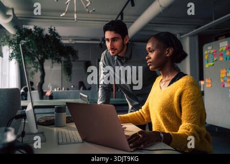 Young male programmer standing beside female colleague using desktop PC in office Stock Photo