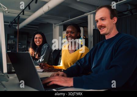 Smiling businesswomen sitting with male colleague using laptop while working in tech office Stock Photo