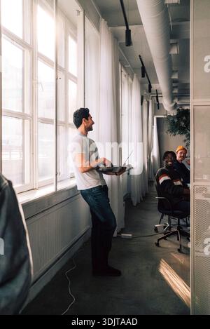 Young male programmer holding laptop talking with colleagues while standing near window in office Stock Photo