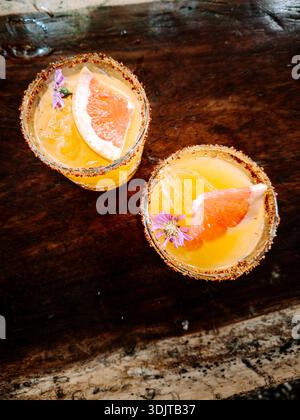 Top-down view of two fresh grapefruit cocktails with chili salt rims, edible flowers, and ice, served on a rustic wooden table in Tulum, Mexico. Stock Photo
