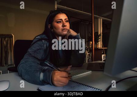 Thoughtful female business professional writing on diary while looking at computer in startup office Stock Photo