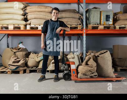 Smiling warehouse worker leaning on a manual forklift trolley in a storage facility. Young cheerful man wearing an apron relaxes during a warehouse sh Stock Photo