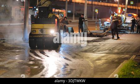 Road roller compacting fresh asphalt during evening roadworks in city street Stock Photo