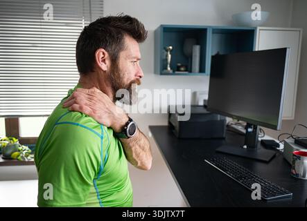Man sitting at a desk in home office holding his neck and experiencing pain while working on a computer. Concept of neck pain, back pain and workplace Stock Photo