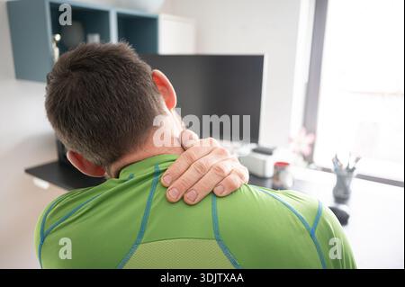 Man sitting at a desk in home office holding his neck and experiencing pain while working on a computer. Concept of neck pain, back pain and workplace Stock Photo