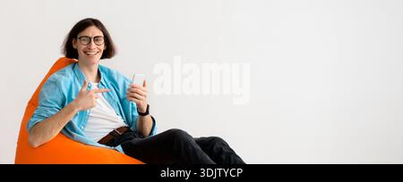 Young Man Sits on Orange Bean Bag While Using Smartphone and Smiling Indoors Stock Photo