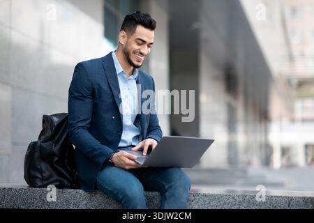 Positive middle eastern guy sitting on the street, using laptop Stock Photo