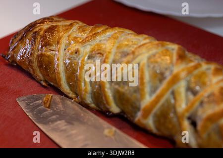 Golden brown beef wellington pastry-wrapped meat on a red cutting board with a knife Stock Photo