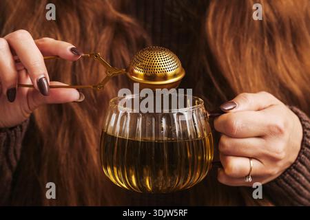 Woman preparing loose leaf tea using metal infuser and glass teapot. Herbal tea brewing ritual at home. Warm relaxing beverage preparation. Stock Photo