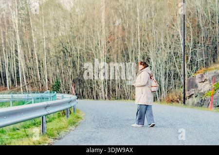 Female traveler in beige coat stands on winding road, surrounded by trees, using smartphone to capture moments, illustrating outdoor exploration and c Stock Photo