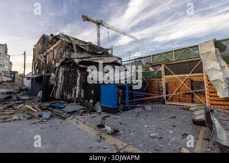 A heavily damaged building shows signs of fire and explosion collapsed roof charred walls scattered debris and a blue industrial unit wrapped in cauti Stock Photo
