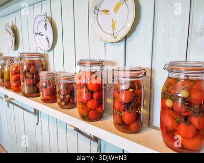Jars filled with preserved tomatoes and vegetables are arranged on a wooden shelf, with decorative plates mounted on a light blue wall above Stock Photo