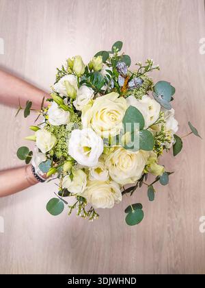 Floral bouquet with white roses and lisianthus, held by hands, surrounded by greenery, set against a light wooden surface Stock Photo