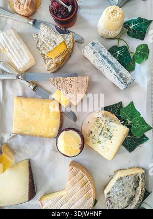 Assorted cheese platter featuring different French cheese varieties, knives, and jars of jam placed on a white tablecloth with ivy leaves for decorati Stock Photo