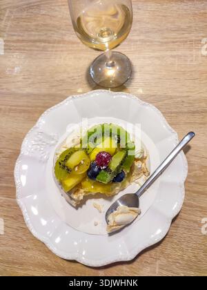 Meringue dessert topped with kiwi, pineapple, and berries on a decorative plate, accompanied by a glass of white wine on a wooden surface Stock Photo