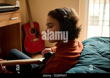 A teen boy in an orange hoodie sits on a bed, wearing headphones, playing a guitar in a sunlit bedroom with a red acoustic guitar nearby. Stock Photo