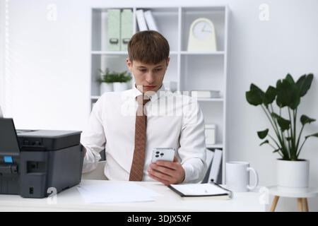 Man using printer and smartphone at white desk in office Stock Photo