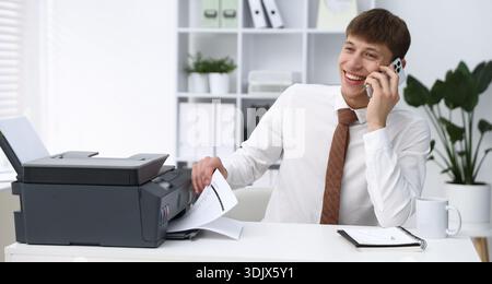 Smiling man taking printed sheet of paper from printer and talking by smartphone at white desk in office Stock Photo