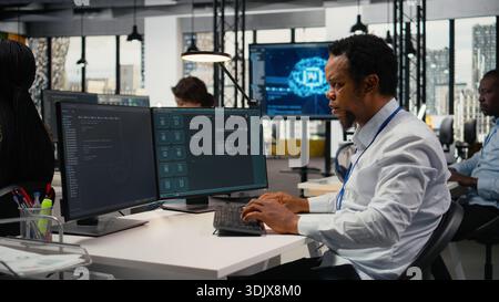 Technician in office solving tasks with artificial intelligence LLM chatbot answering prompts. African american man at work using PC to interact with large language model program, camera A Stock Photo