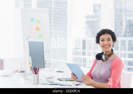 Woman sitting at desk near windows holding tablet, facing camera, monitor sticky notes, copy space. Minimalist, modern, bright, sunlit, productivity, Stock Photo