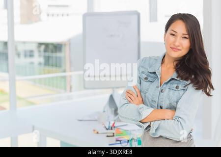 Asian woman standing right in office wearing denim jacket arms crossed near whiteboard, copy space. Professional, confident, modern, bright, windows, Stock Photo