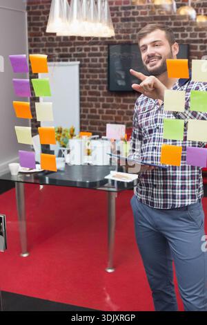 Male standing at glass wall pointing to sticky notes holding tablet in modern office, copy space. Brainstorming, ideation, collaboration, planning, wo Stock Photo