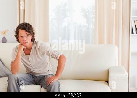 Man sitting on white leather sofa in living room, wearing beige henley and looking toward camera. Person, couch, interior, minimal, neutral, daylight, Stock Photo