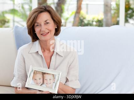 Senior woman sitting on sofa holding silver framed picture of two children near windows, copy space. Grandmother, portrait, cozy, livingroom, cushion, Stock Photo