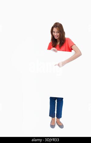 Woman is holding blank white rectangular board and pointing at lower area in studio, copy space. Backdrop, highkey, placard, presenter, casual, minima Stock Photo
