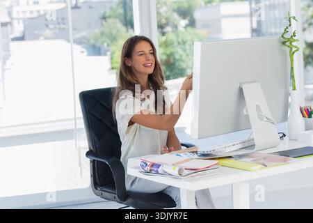 Woman sitting at white desk in modern office, working on desktop monitor with keyboard and mouse. Professional, workspace, daylight, urban, plant, min Stock Photo