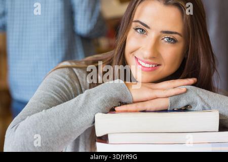 Woman leaning on stack of thick textbooks, smiling at camera in library wearing grey sweater. Student, books, study, education, learning, focus, portr Stock Photo
