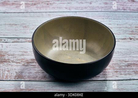 A striking empty ceramic bowl, featuring a rough dark exterior and a speckled beige-green interior, sits centered on a bright, rustic wooden tabletop. Stock Photo