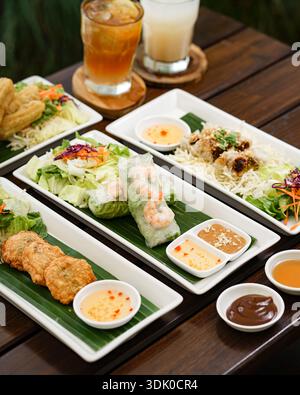 Delicious Asian dishes including spring rolls, salads, and fried items served on white plates with various dipping sauces and cool beverages on a rust Stock Photo