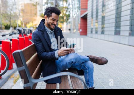 Young adult Latin American man smiling while using smartphone on bench Stock Photo
