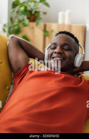 African man lying on sofa listening to music with wireless headphones at home Stock Photo