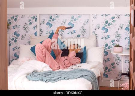 Wrapped in a soft pink onesie, a young woman enjoys a peaceful moment lounging on her bed. Surrounded by floral wallpaper, she smiles while scrolling Stock Photo